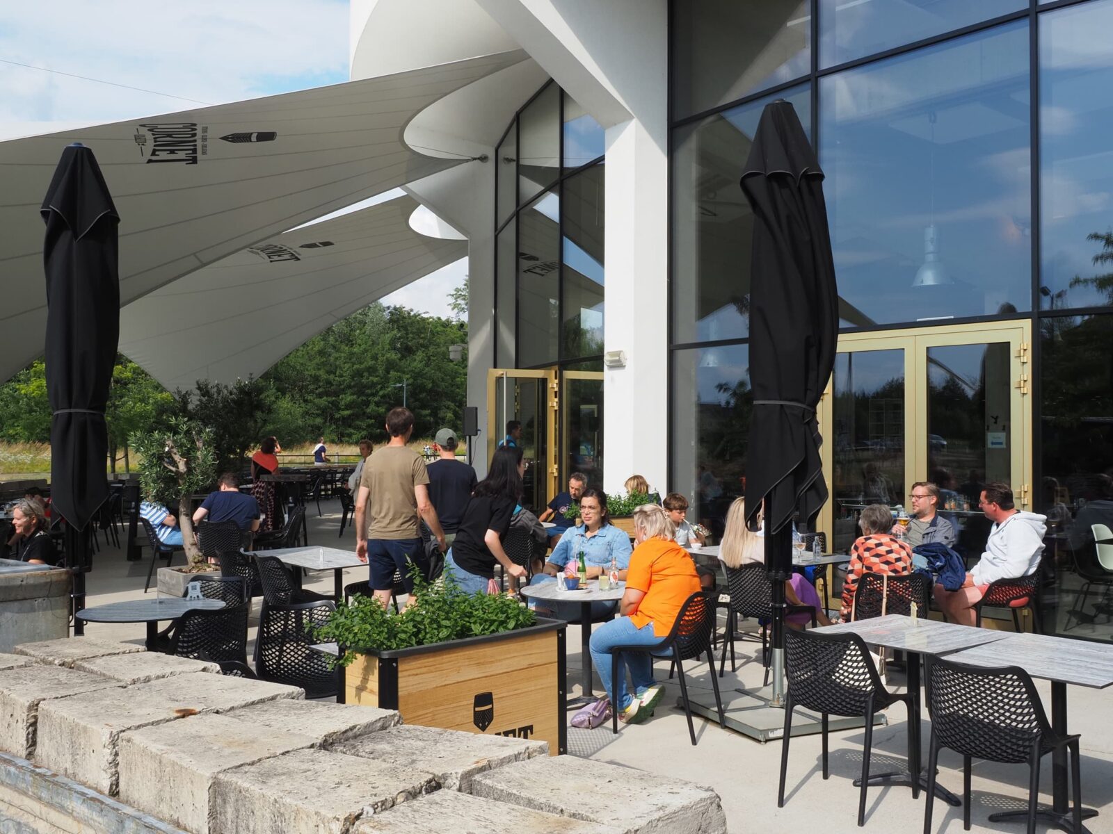 Bezoekers zitten met een drankje op het terras van de TODI-brasserie, onder grote schaduwdoeken.